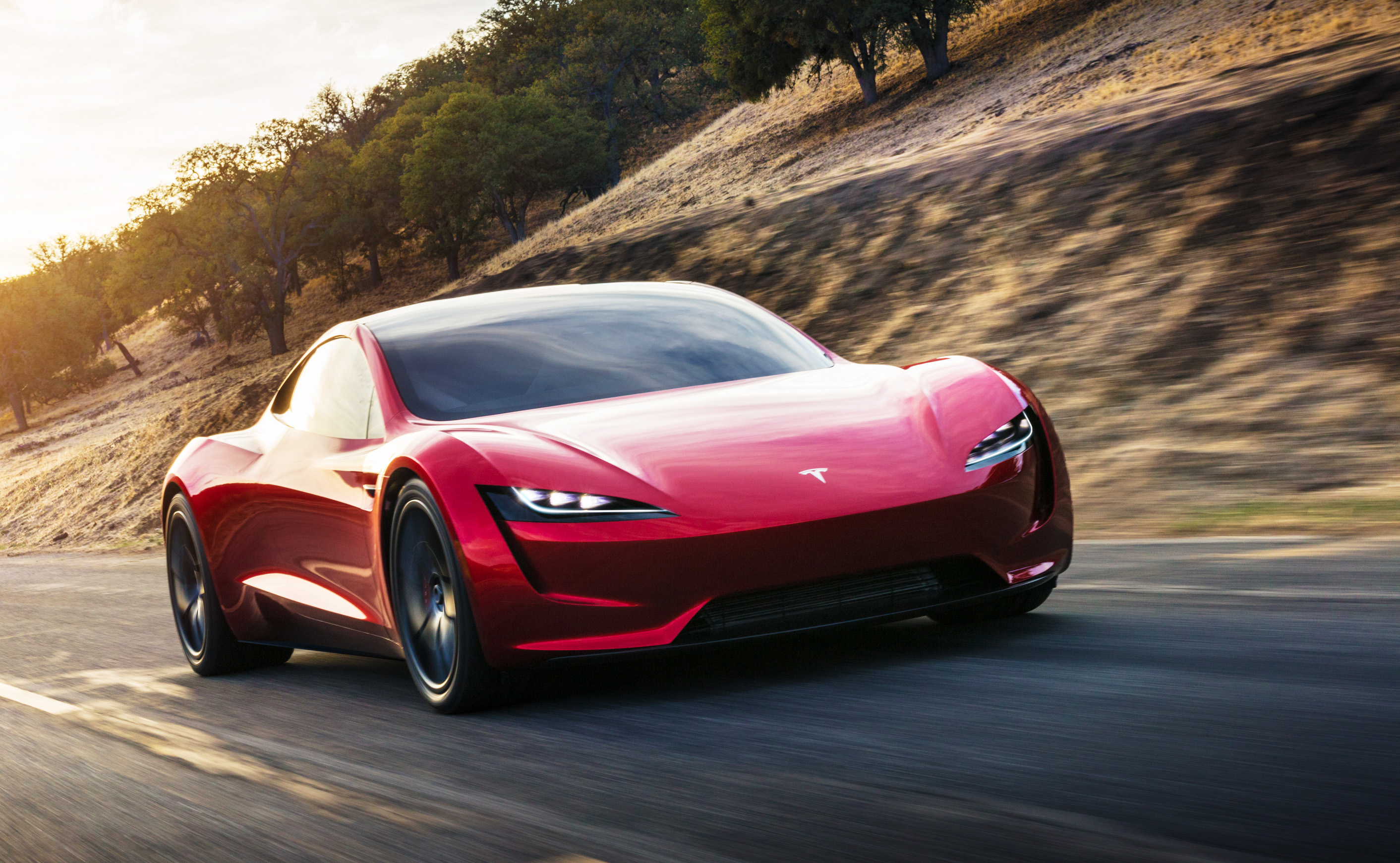 Red Tesla Roadster on the road with a dirt hill and trees in the background. (Custard, Ben. All New 2022 Tesla Roadster Will Get Plaid Powertrain . Carbuyer UK. 2021, Feb 1.)